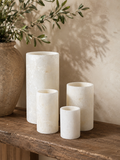 Three cylindrical white stone vases in different heights are arranged on a rustic wooden shelf next to a large textured ceramic pot with green branches, against a warm beige wall with soft leaf shadows.