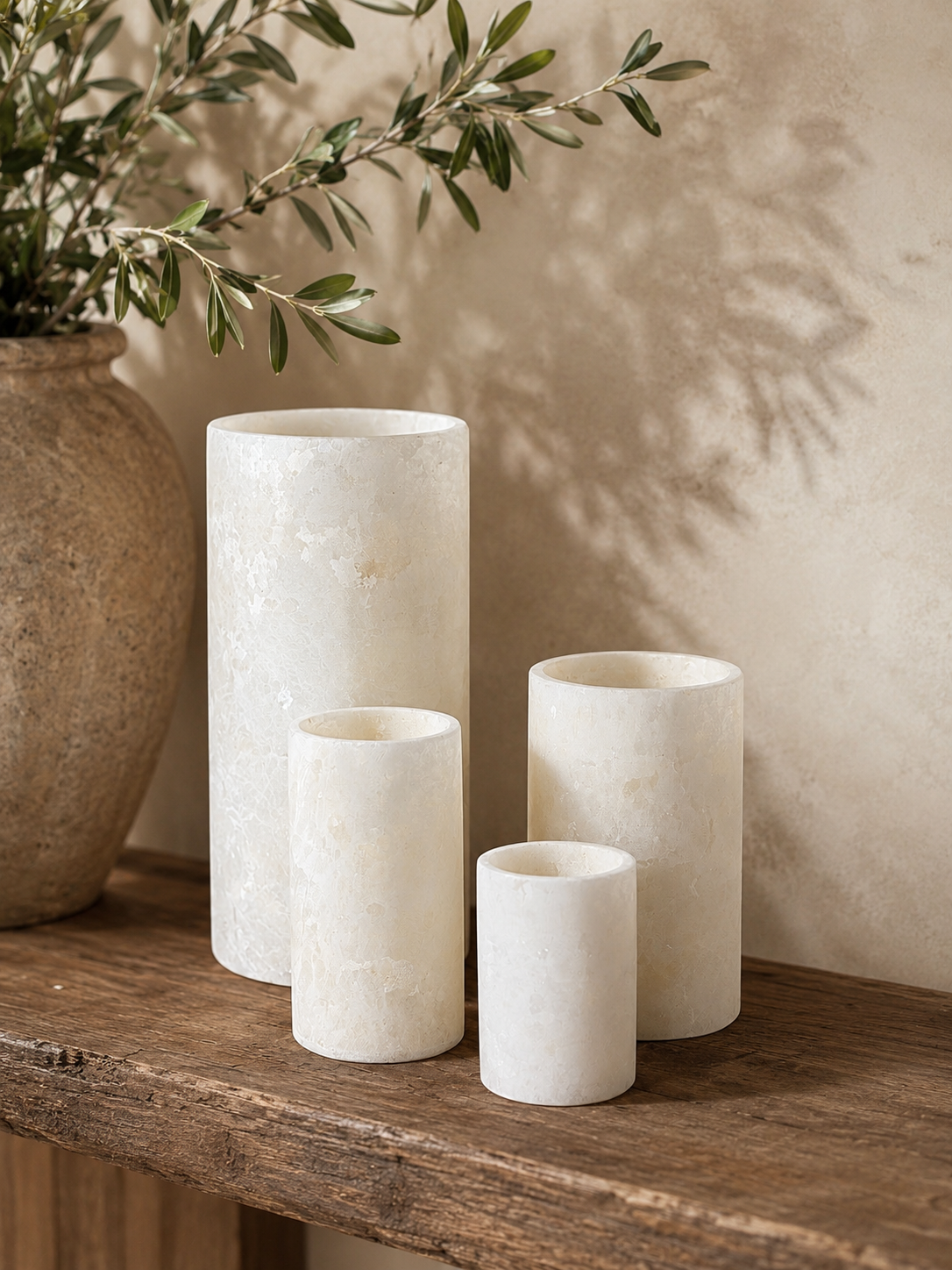 Three cylindrical white stone vases in different heights are arranged on a rustic wooden shelf next to a large textured ceramic pot with green branches, against a warm beige wall with soft leaf shadows.