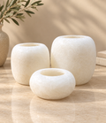 Three round stone candle holders in varying sizes, crafted from a textured cream-colored stone, arranged on a polished beige marble surface with a decorative olive branch in the background.