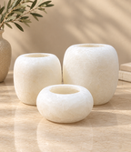 Three round stone candle holders in varying sizes, crafted from a textured cream-colored stone, arranged on a polished beige marble surface with a decorative olive branch in the background.